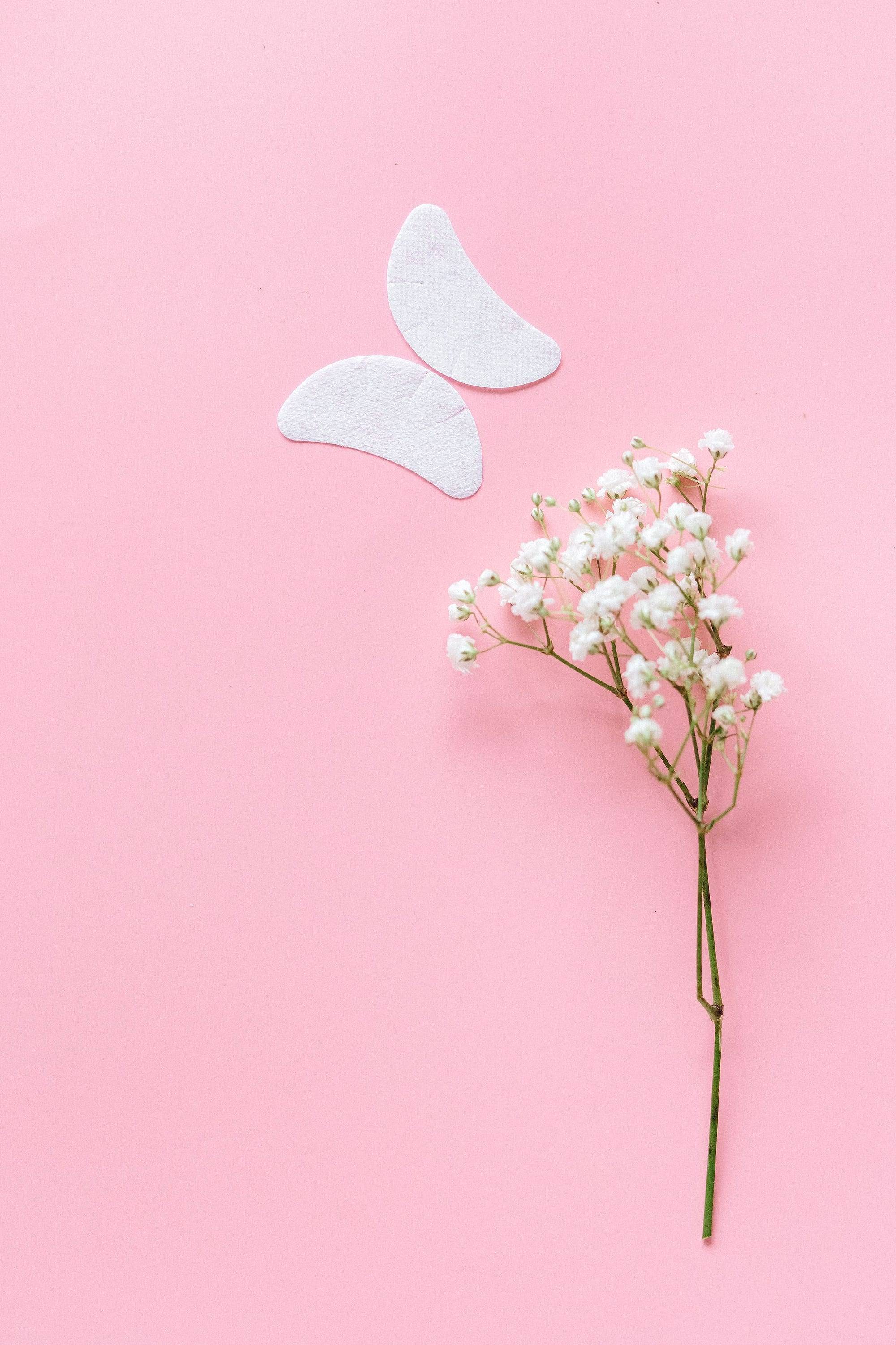 Parches hidratantes para el contorno de ojos junto a una ramita de paniculata sobre fondo rosa