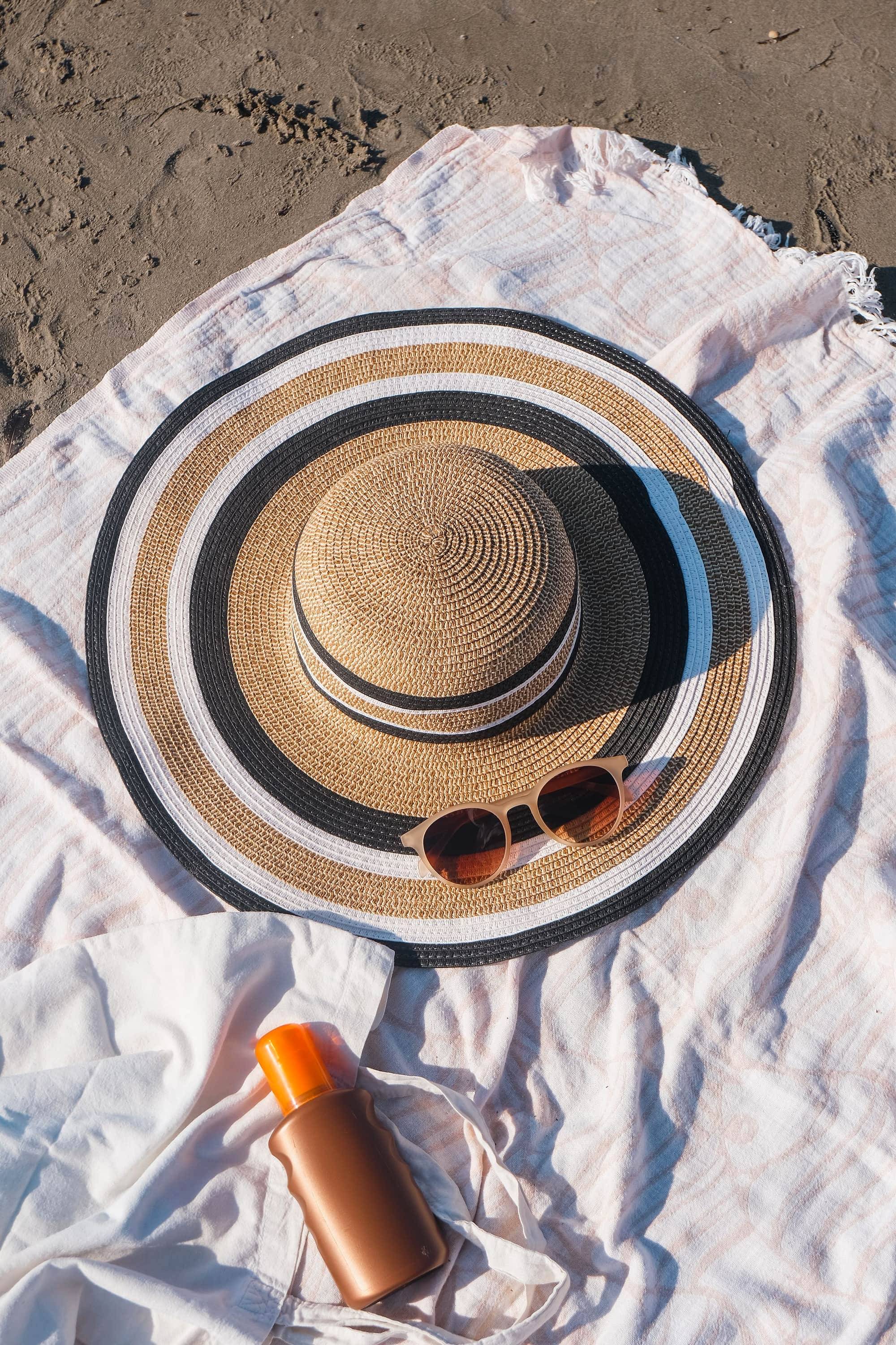 Sombrero de paja, gafas de sol y protector solar sobre una toalla blanca en la playa
