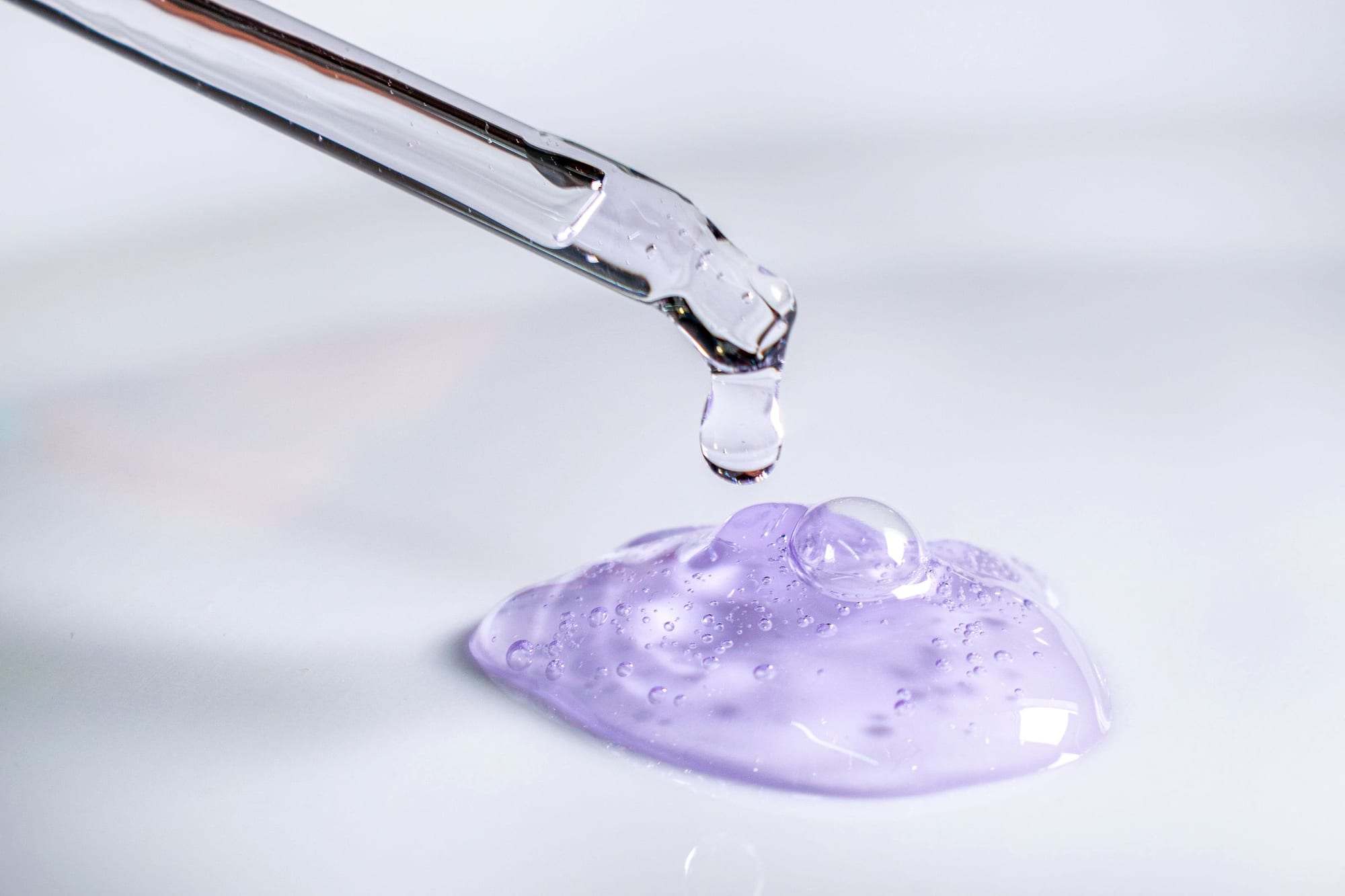 Pipeta dejando caer una gota de gel cosmético lila sobre una superficie blanca
