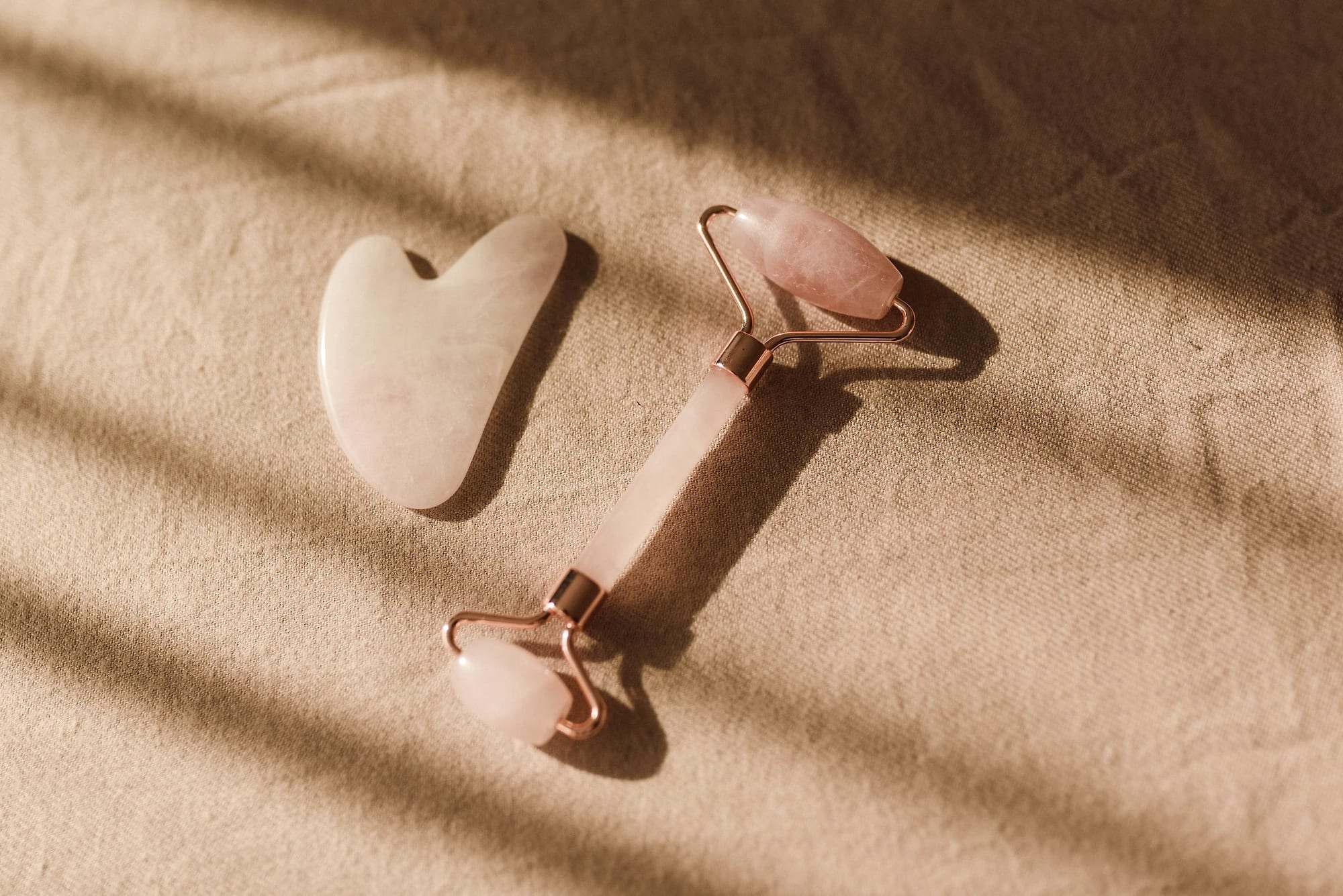 Rodillo facial y piedra gua sha de cuarzo rosa sobre una tela beige con luz suave