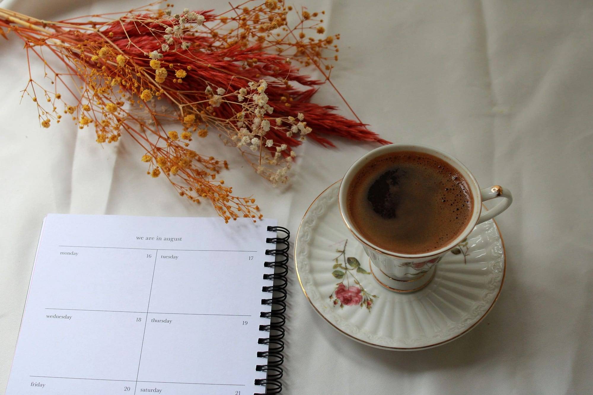 Open planner next to a cup of coffee and dried flowers on light linen