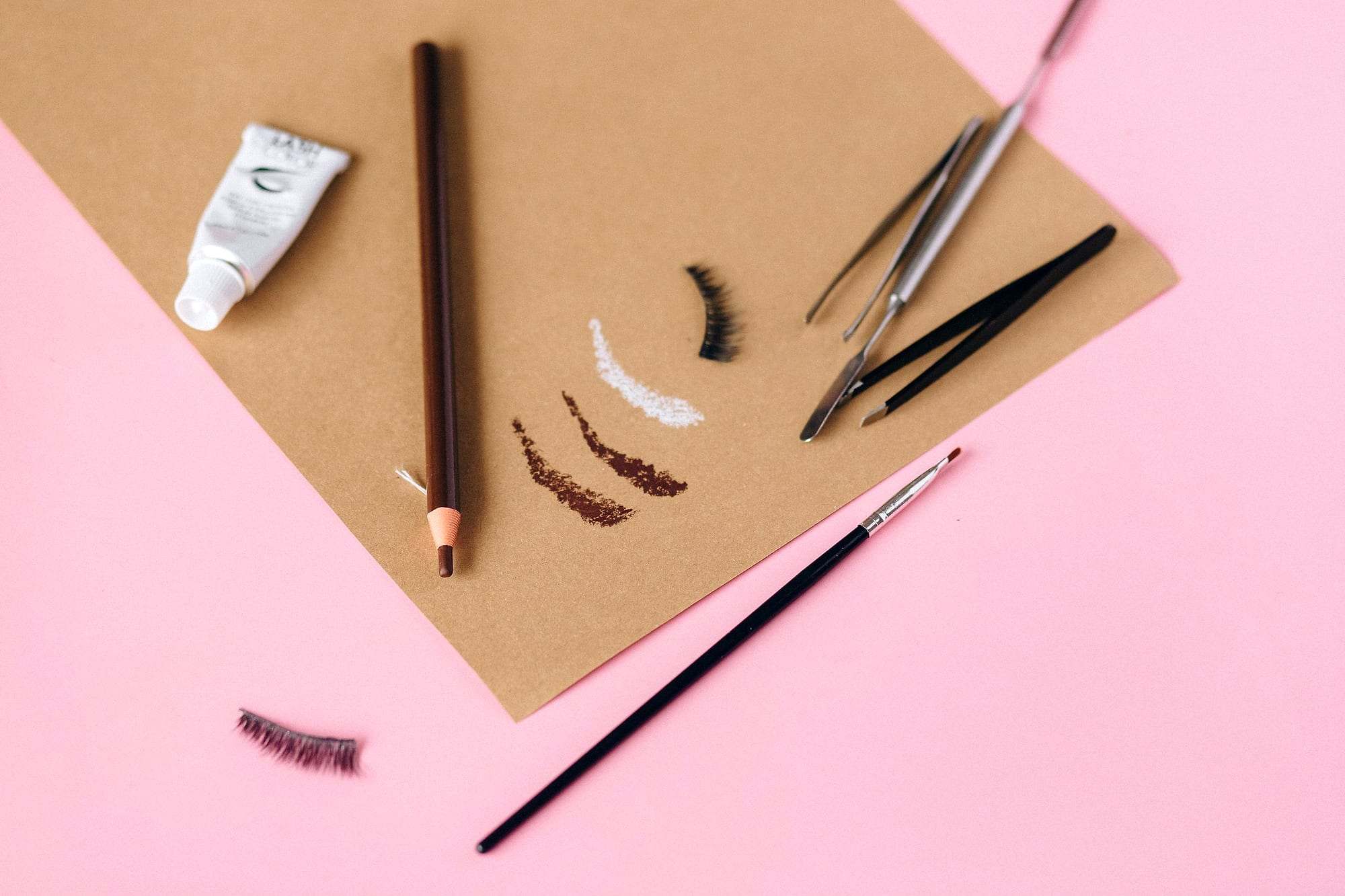 Lápiz de cejas, pinceles, pinzas y muestras de color de cejas sobre papel kraft y fondo rosa